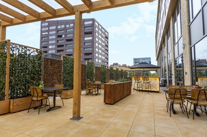 An outdoor patio area with wooden tables and chairs, a central wooden planter, and a backdrop of a tall building.