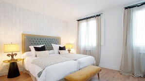 A double bed with a tufted headboard, two bedside tables with lamps, a bench, and a large window with sheer curtains.