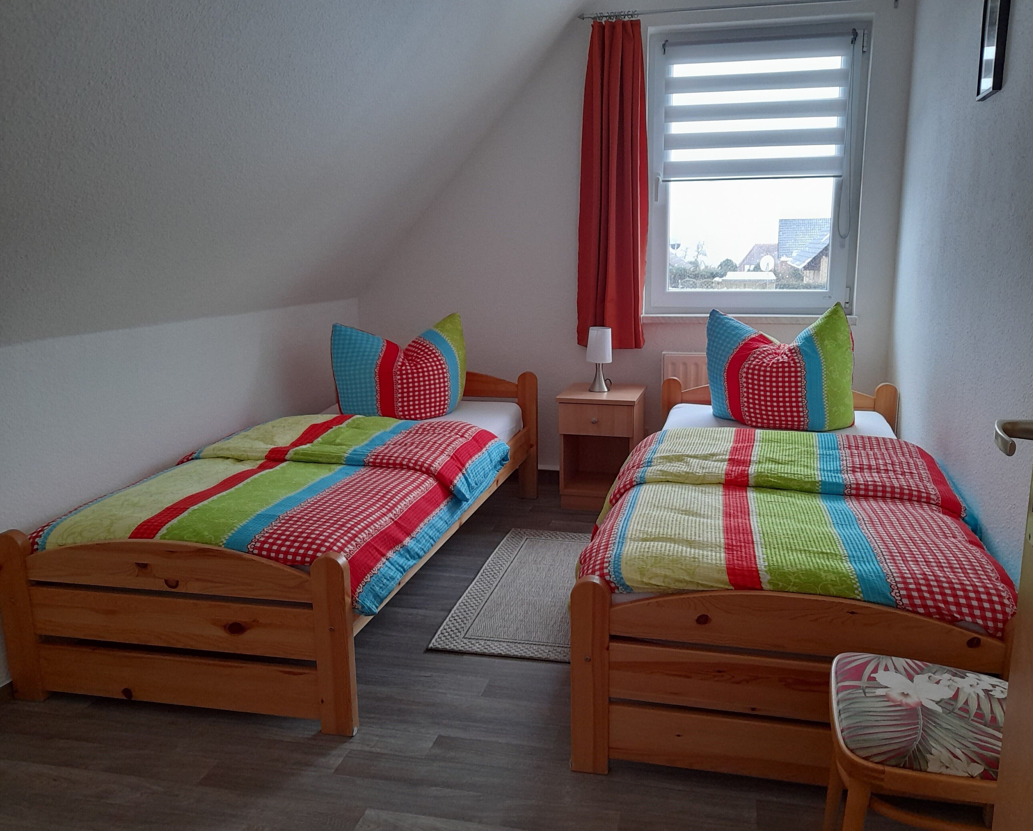 Un dormitorio con dos camas de madera, cada una con ropa de cama y almohadas coloridas. Hay una ventana con persianas, una mesita de noche con lámpara y una silla con cojín estampado.