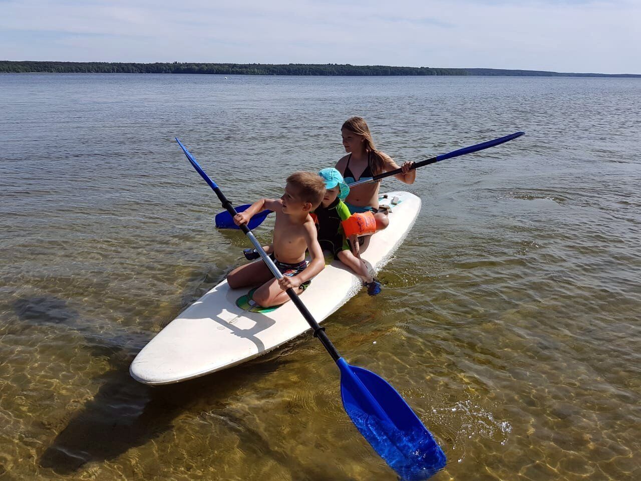 A kayak on a calm lake with clear water, two paddlers, and a distant shoreline.