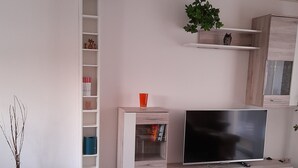 Una sala de estar moderna con un televisor sobre un mueble de madera, una estantería y una planta en un estante.