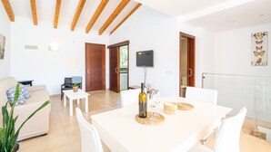 A modern living room with a white sofa, a dining table with chairs, a TV mounted on the wall, and a wooden ceiling. - Apartment Cala Anguilla  (Manacor Cala Anguila)