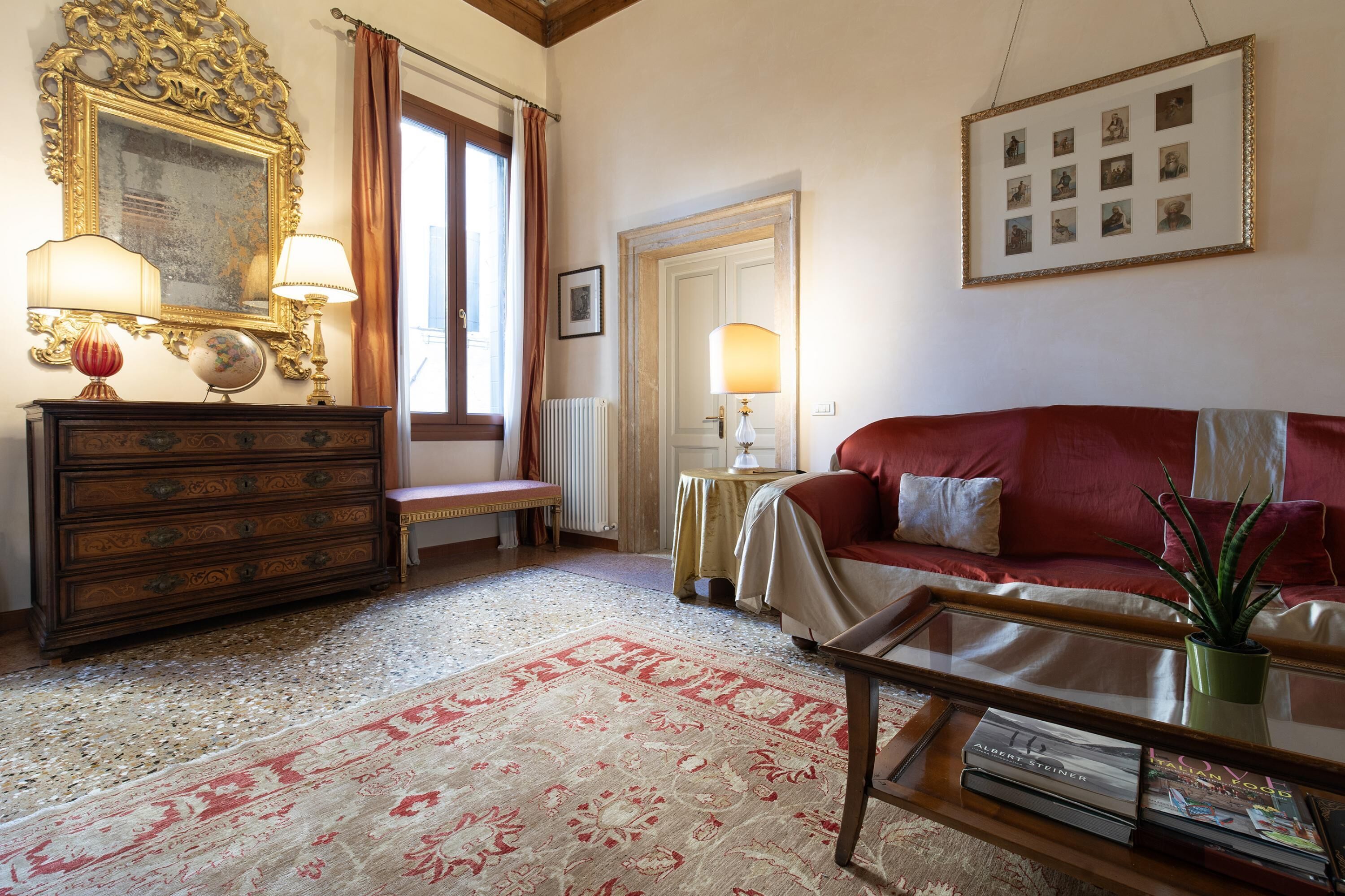 Un salon avec un canapé rouge, une commode en bois, un globe terrestre et un mur orné d’un ensemble de photos encadrées.