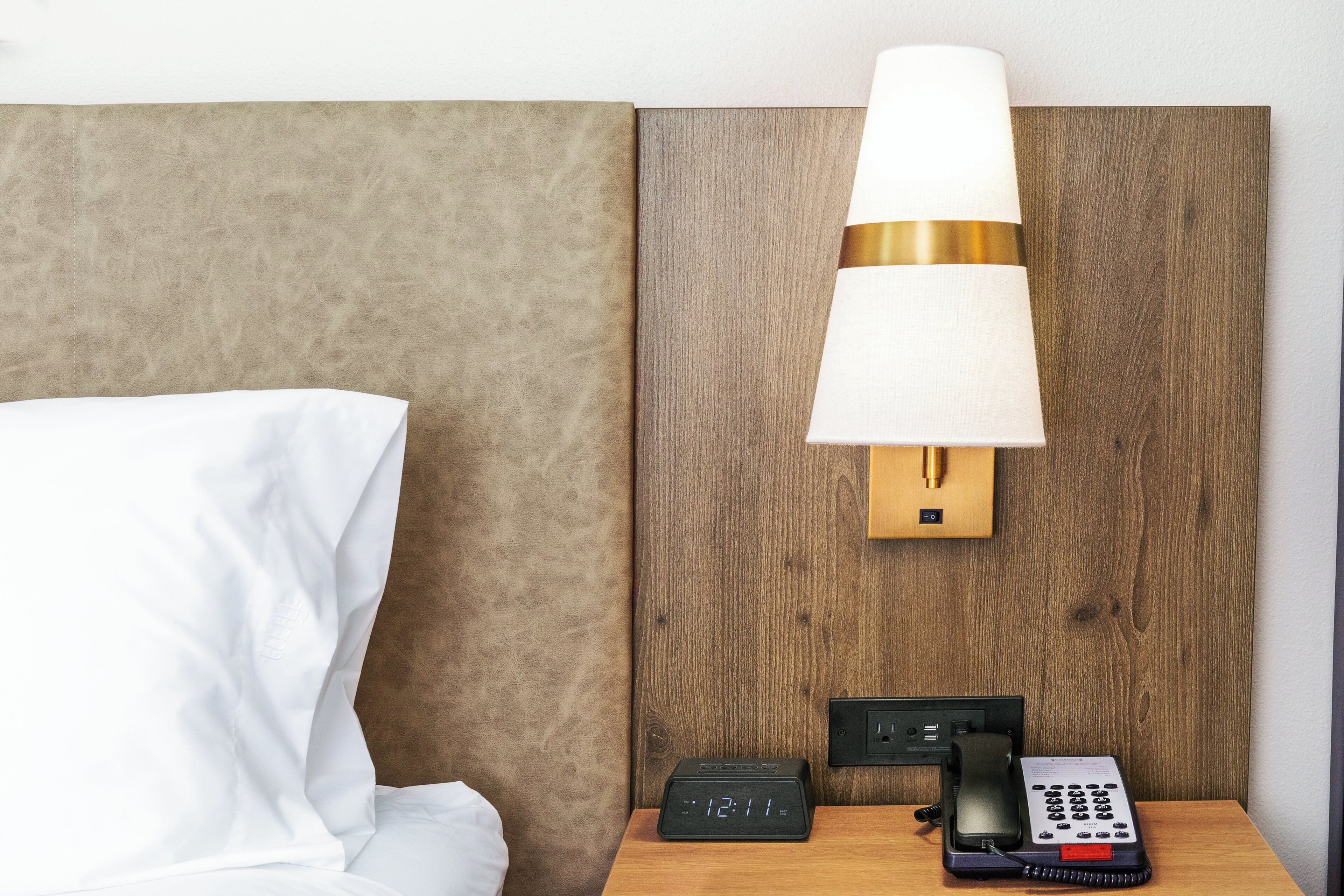 Una habitación de hotel con una cama, una mesita de noche con lámpara, un reloj y un teléfono.