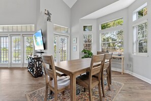 Un vaste espace dédié à la salle à manger, avec une table et des chaises en bois, un téléviseur posé sur un meuble et de grandes fenêtres équipées de stores.