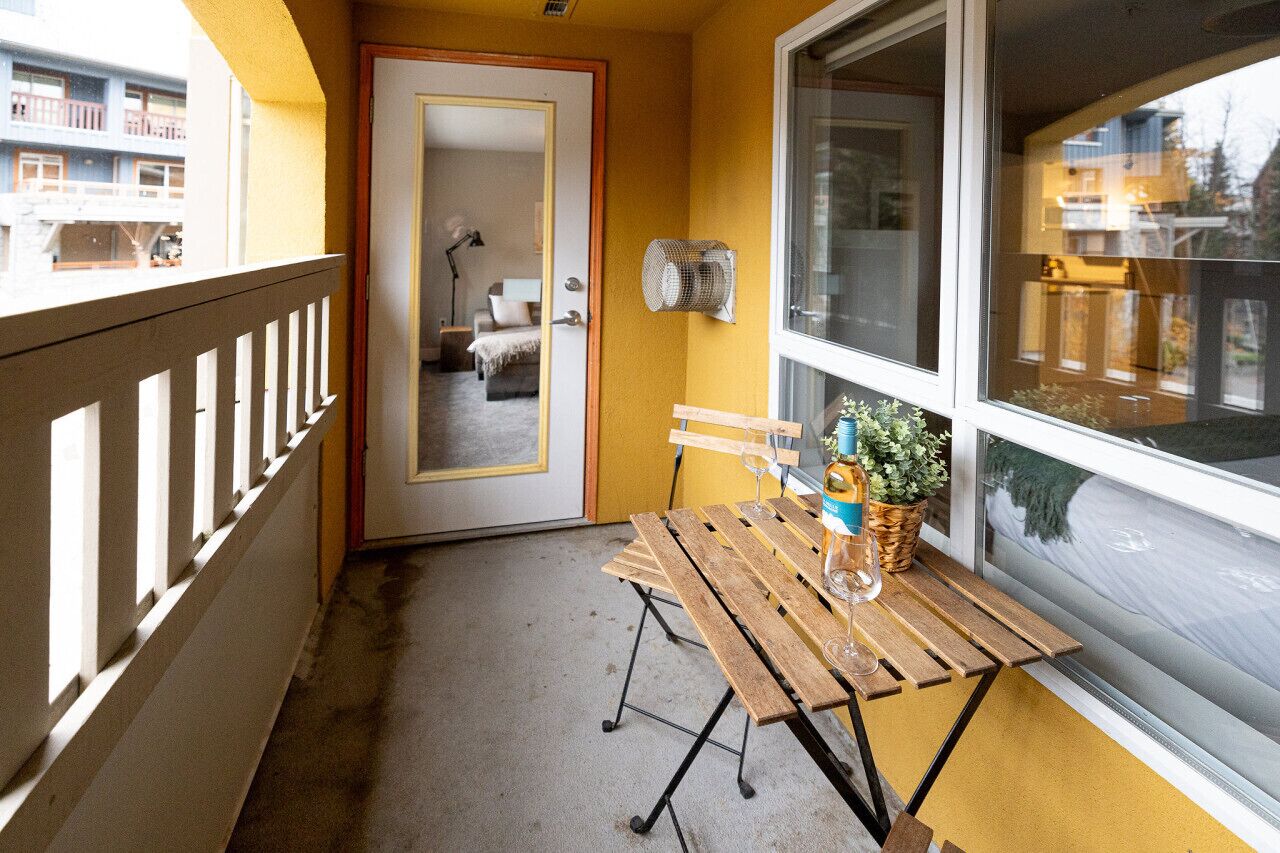 Un balcon avec une table en bois, une chaise, un pot de fleurs et une fenêtre donnant sur des immeubles.