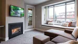A living room with a fireplace, a television mounted on the wall, and a large window with a view of buildings outside. - ❄️ Whistler Center 1BR:Walk Everywhere. Free Parking. Fire, Sleeps 4 ❄️ (Whistler)
