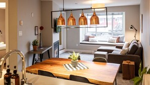 A modern living room with a wooden dining table, a sofa, and a window with a view of buildings. - ❄️ Whistler Center 1BR:Walk Everywhere. Free Parking. Fire, Sleeps 4 ❄️ (Whistler)