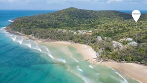 Vue aérienne d’une zone côtière avec une plage, des eaux turquoise et limpides, et une colline boisée.