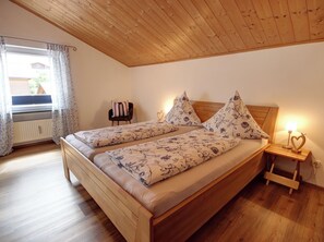 A bedroom with a wooden ceiling, a bed with floral bedding, a chair, a nightstand with a lamp, and a window with curtains.