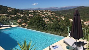 A pool with clear blue water, surrounded by lounge chairs and an umbrella, overlooking a hillside village. - Large stylish apartment with sea views over the Bay of Cannes and infinity pool (Les Adrets-de-l'Estérel)