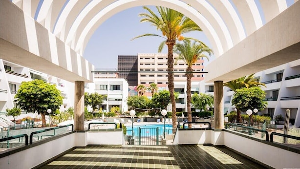 A modern hotel atrium with a pool, palm trees, and a clear sky.
