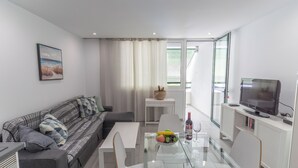 A modern living room with a sofa, a glass dining table with fruit, a TV, and a balcony with curtains.