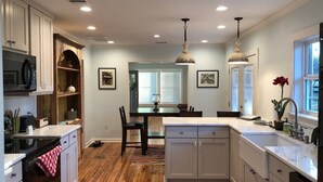 Una cocina con suelo de madera, armarios blancos, una isla central, iluminación colgante y una zona de comedor con sillas y mesa.