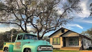 Una camioneta verde antigua estacionada frente a una casa pequeña con techo inclinado y un porche.