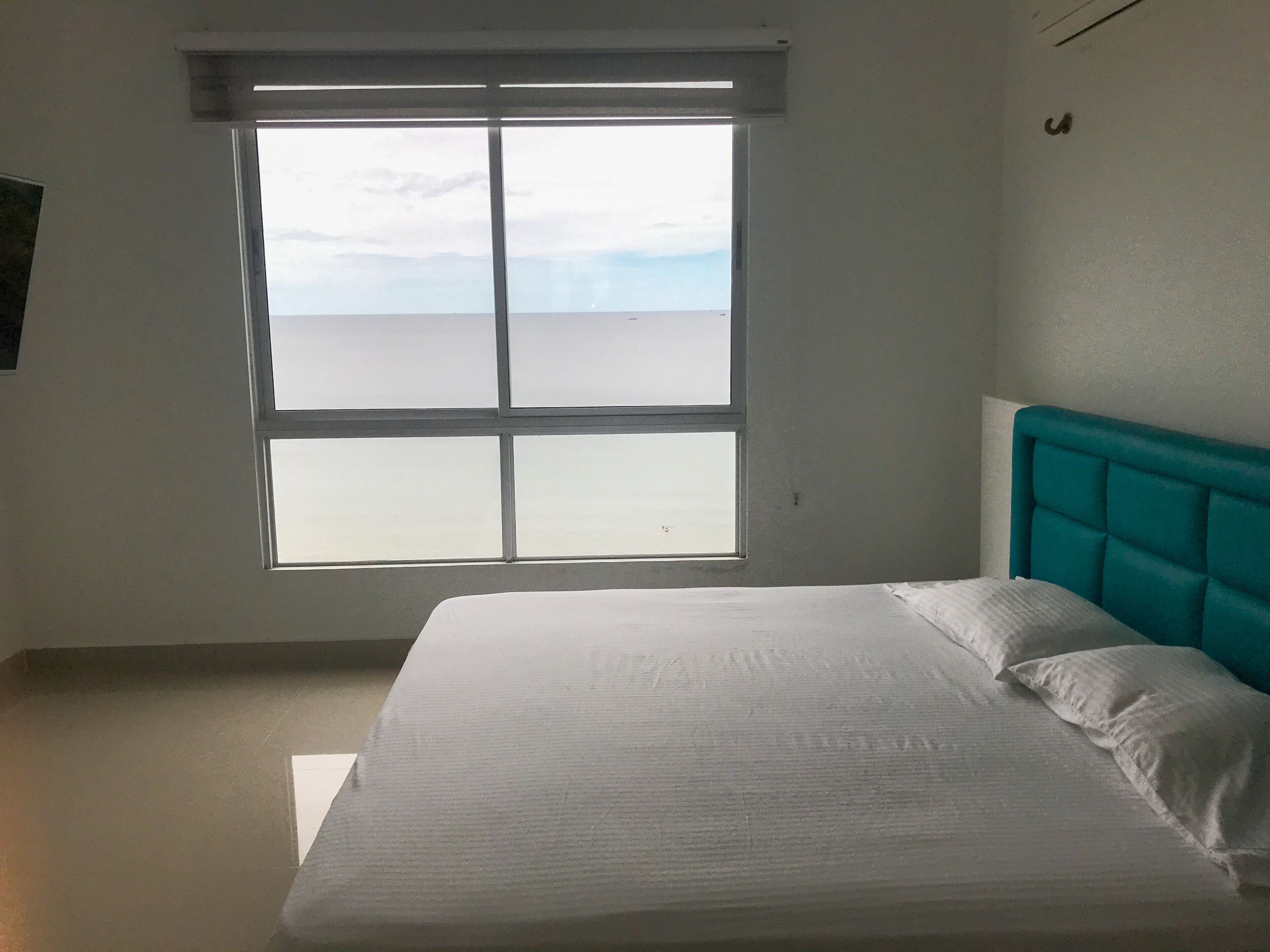 Ein Zimmer mit großem Fenster, einem Bett mit weißer Bettwäsche und einem türkisblauen Kopfteil.