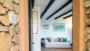 Pared de piedra con un letrero con el número '93', una puerta blanca, un sofá blanco con cojines verdes y grises, y una obra de arte floral enmarcada en la pared.