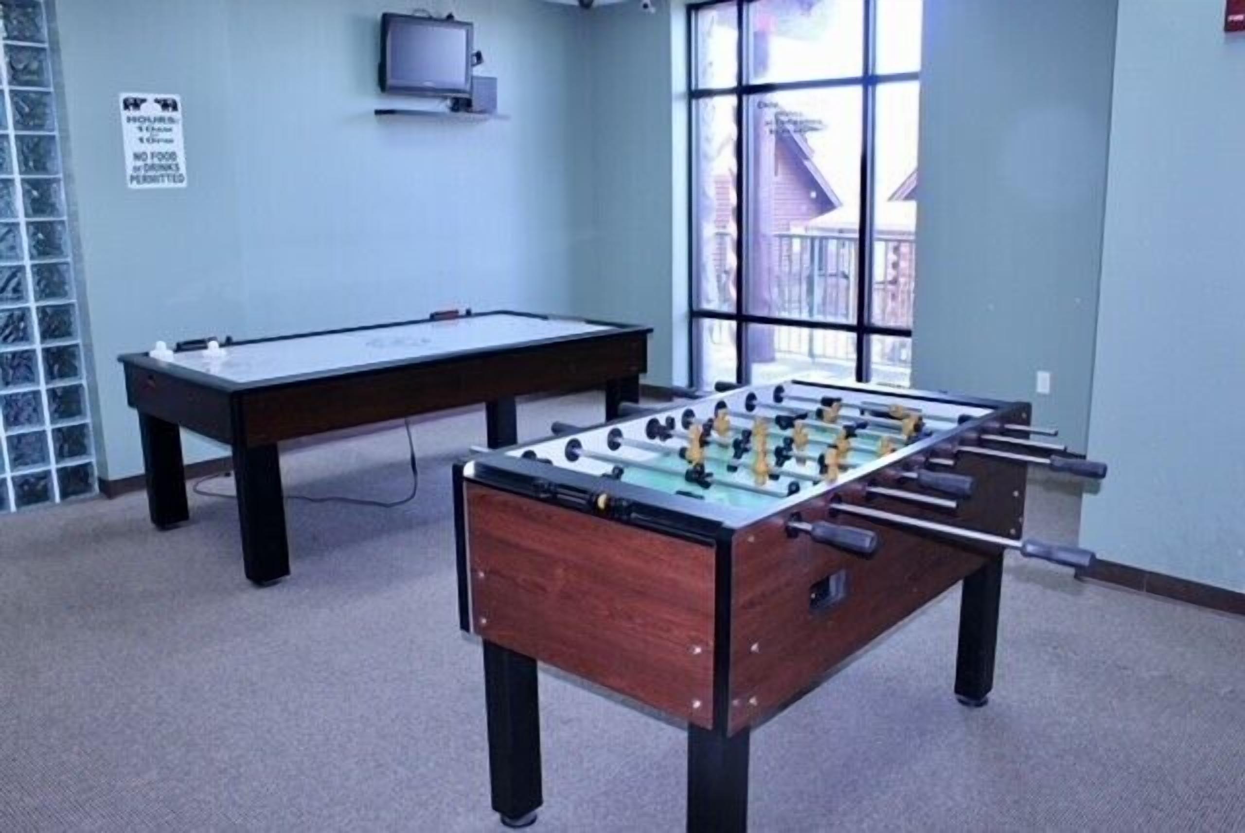 A room with a foosball table, a pool table, and a television on the wall.