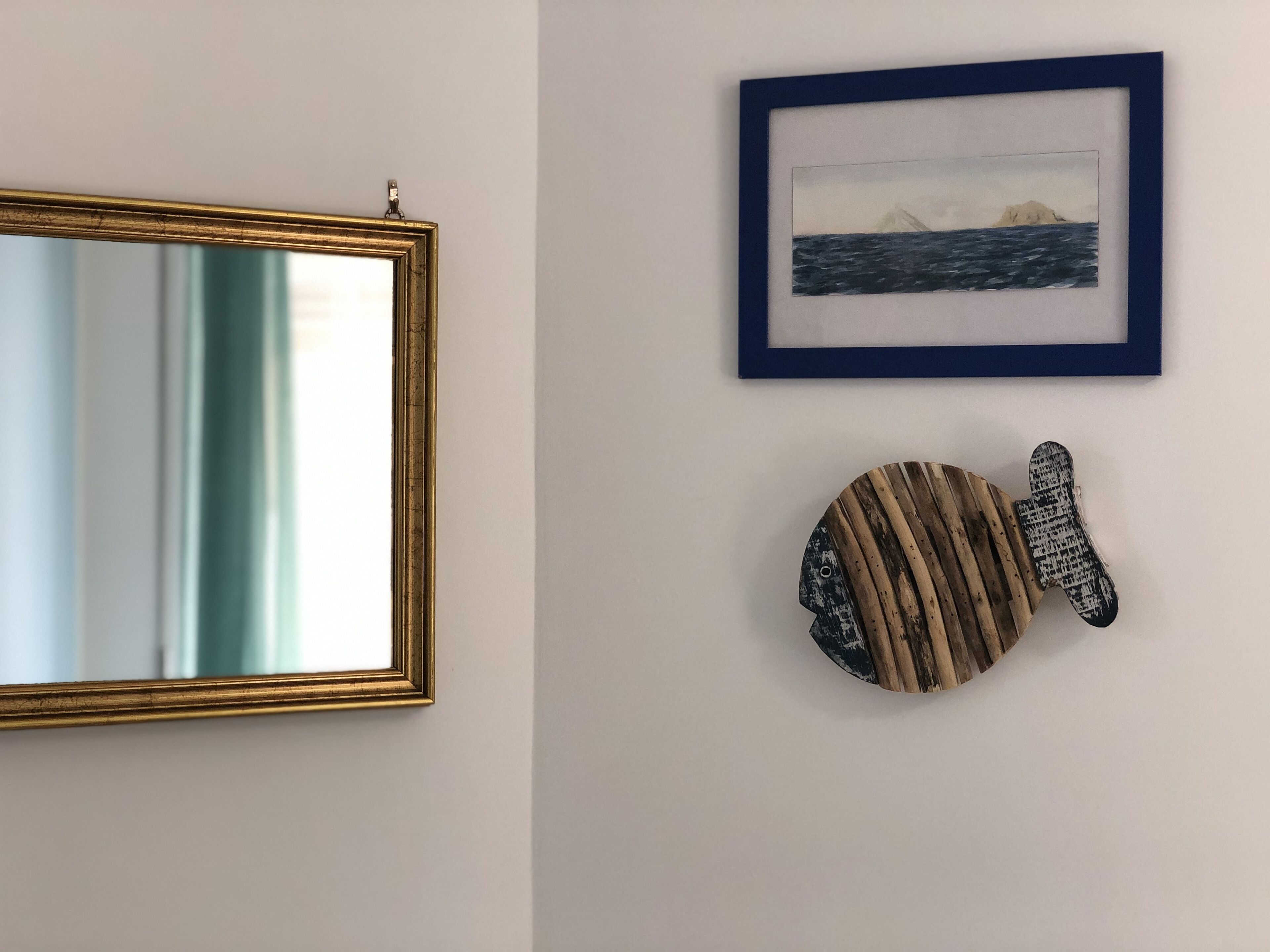 A wall with a framed picture of a landscape, a mirror, and a decorative fish sculpture.