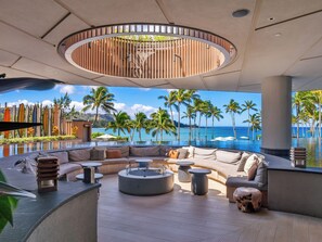 Un área lounge moderna con una disposición de asientos curvada, una fogata en el centro y vistas al océano y palmeras.