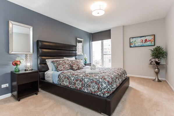 A modern bedroom with a dark headboard, a patterned bedspread, a nightstand with a vase of flowers, and a large window offering a city view. - MAG MILE MEGA Entire Floor 4BD 4BA +Rooftop&Gym (Chicago)