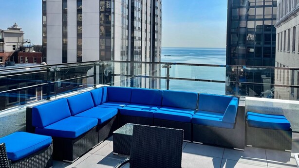 A rooftop terrace with a blue sectional sofa and wicker chairs, overlooking a cityscape and the ocean.