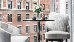 A modern room with a view of a brick building, a round table with a vase of flowers, and two upholstered chairs.