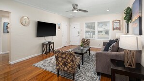 Una sala de estar con piso de madera, ventilador de techo, televisor de pantalla plana, una mesa de centro, dos sillones estampados, un sofá, un tapete y un reloj fijado a la pared.