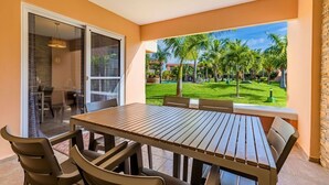Une terrasse avec une table et des chaises en bois, une porte vitrée donnant sur une pièce, et une vue sur une pelouse bien entretenue agrémentée de palmiers.