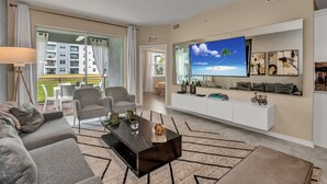 A modern living room with a large flat-screen TV, a sofa, a coffee table, and a view of a balcony with outdoor furniture. - 3180paraunit101 (Kissimmee)