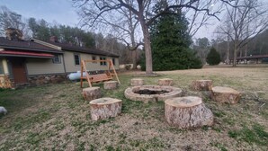 A backyard with a fire pit surrounded by tree stump seating, a swing set, and a house in the background. - Eagle's Nest by Table Rock. Perfect for Hickers, Cyclers, and Swimmers. (Sunset)