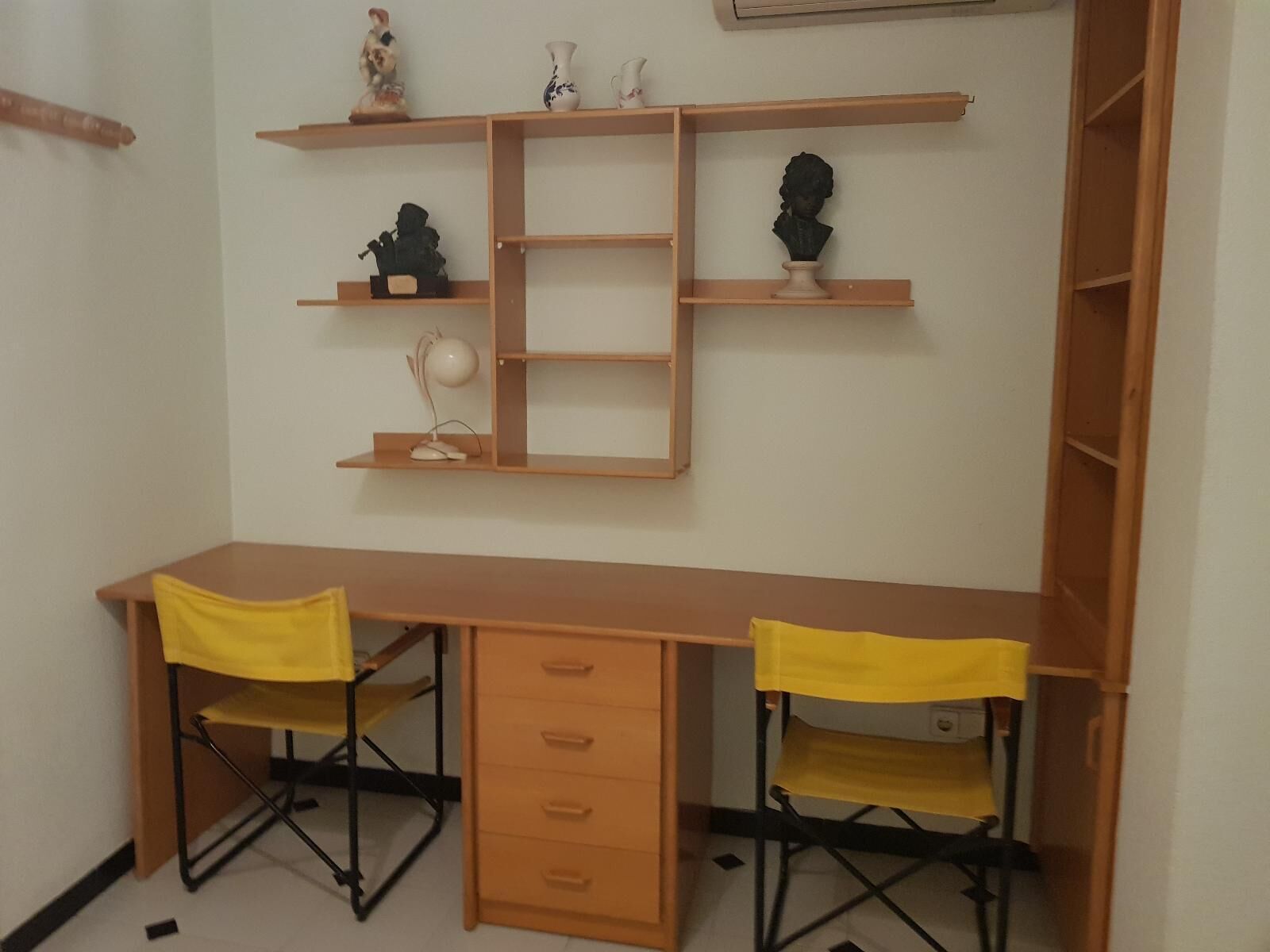 A study room with a wooden desk, two yellow chairs, a lamp, and shelves with decorative items.
