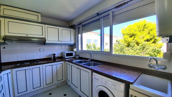 A kitchen with white cabinets, a microwave, a washing machine, and a window with a view of trees and buildings. - 140m2 apartment with lateral sea views D6 (Gandia)
