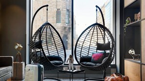 A room with two hanging chairs, a small table, a suitcase, a leather bag, and a view of a cityscape through large windows. - Moxy Edinburgh (Edinburgh)