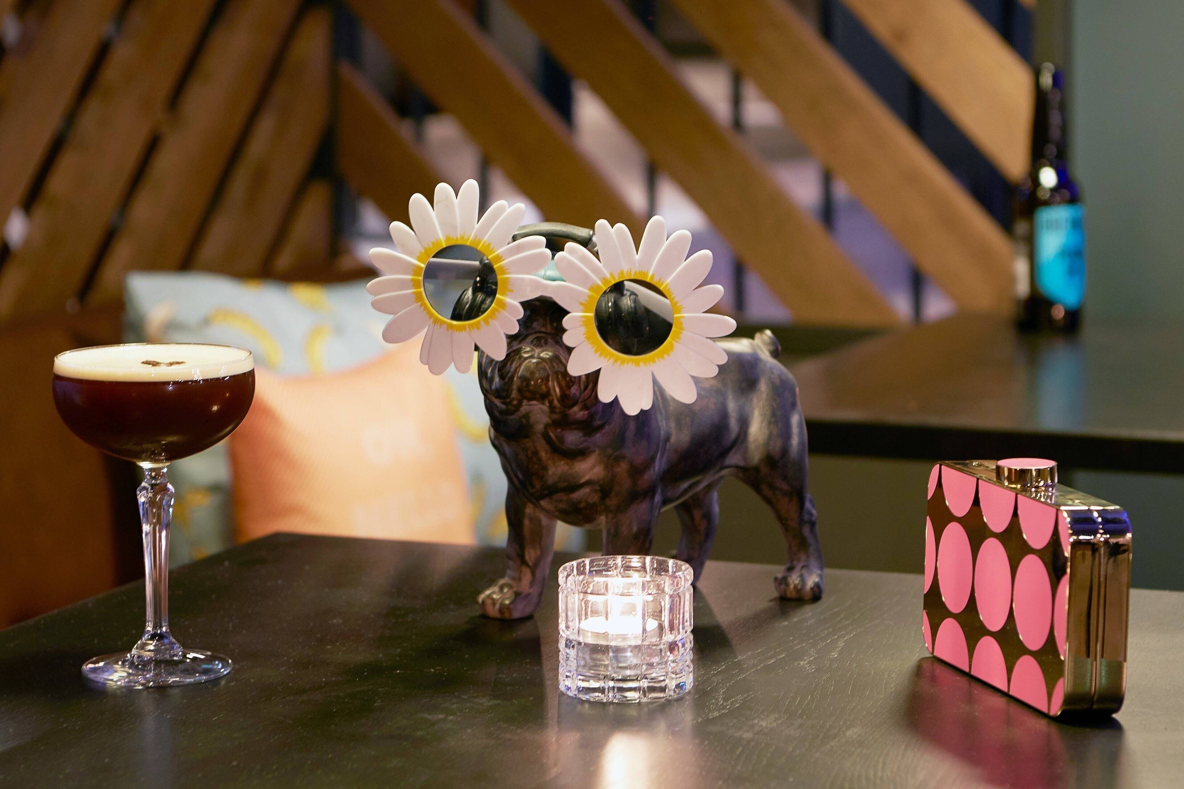 Una mesa con una copa de vino tinto, una vela pequeña, una figurita decorativa de un perro y un encendedor con estampado rosa y negro.