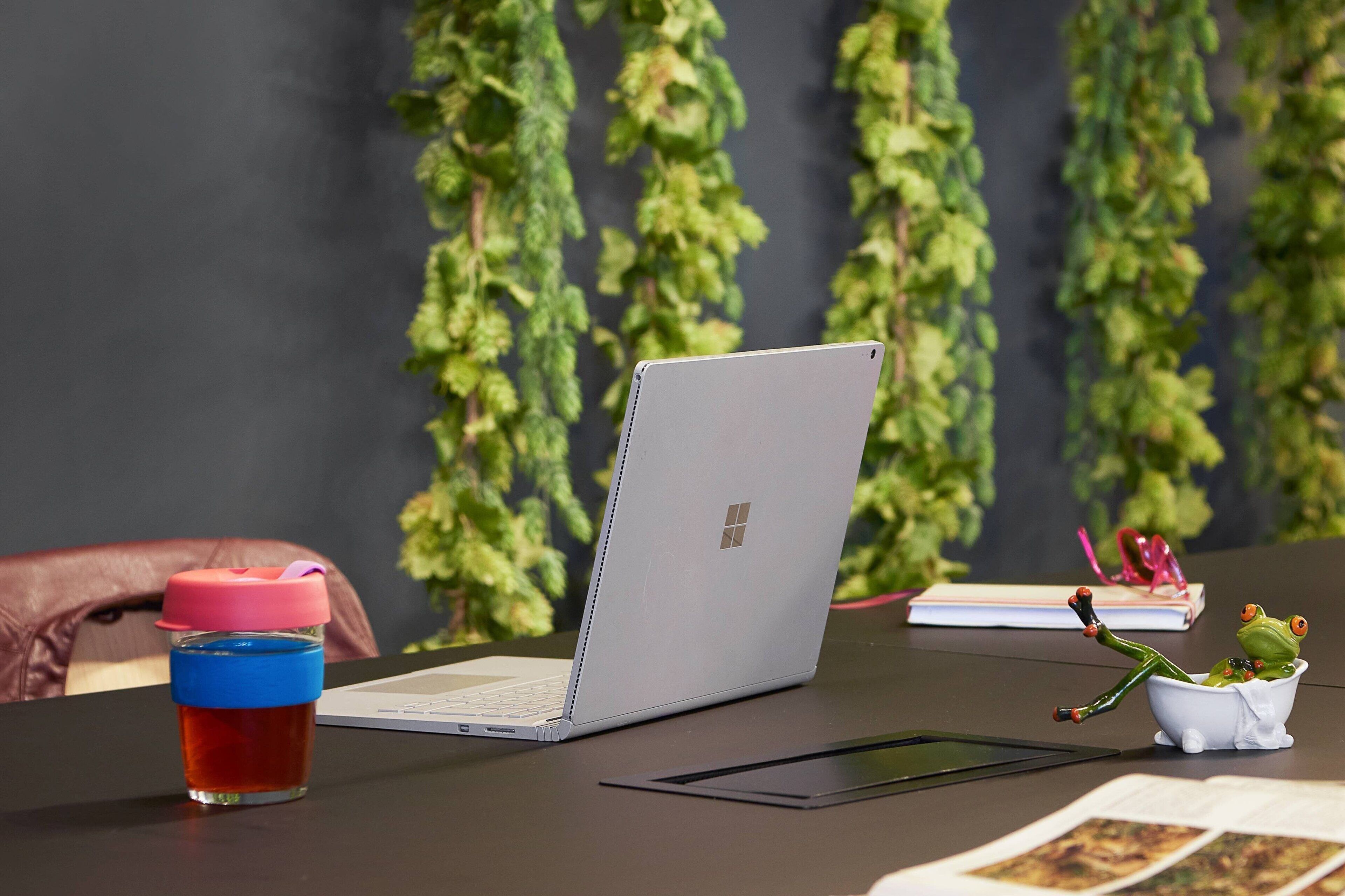 Una computadora portátil sobre una mesa con una pequeña figurina de un sapo en una bañera, una taza con tapa y un libro al fondo.