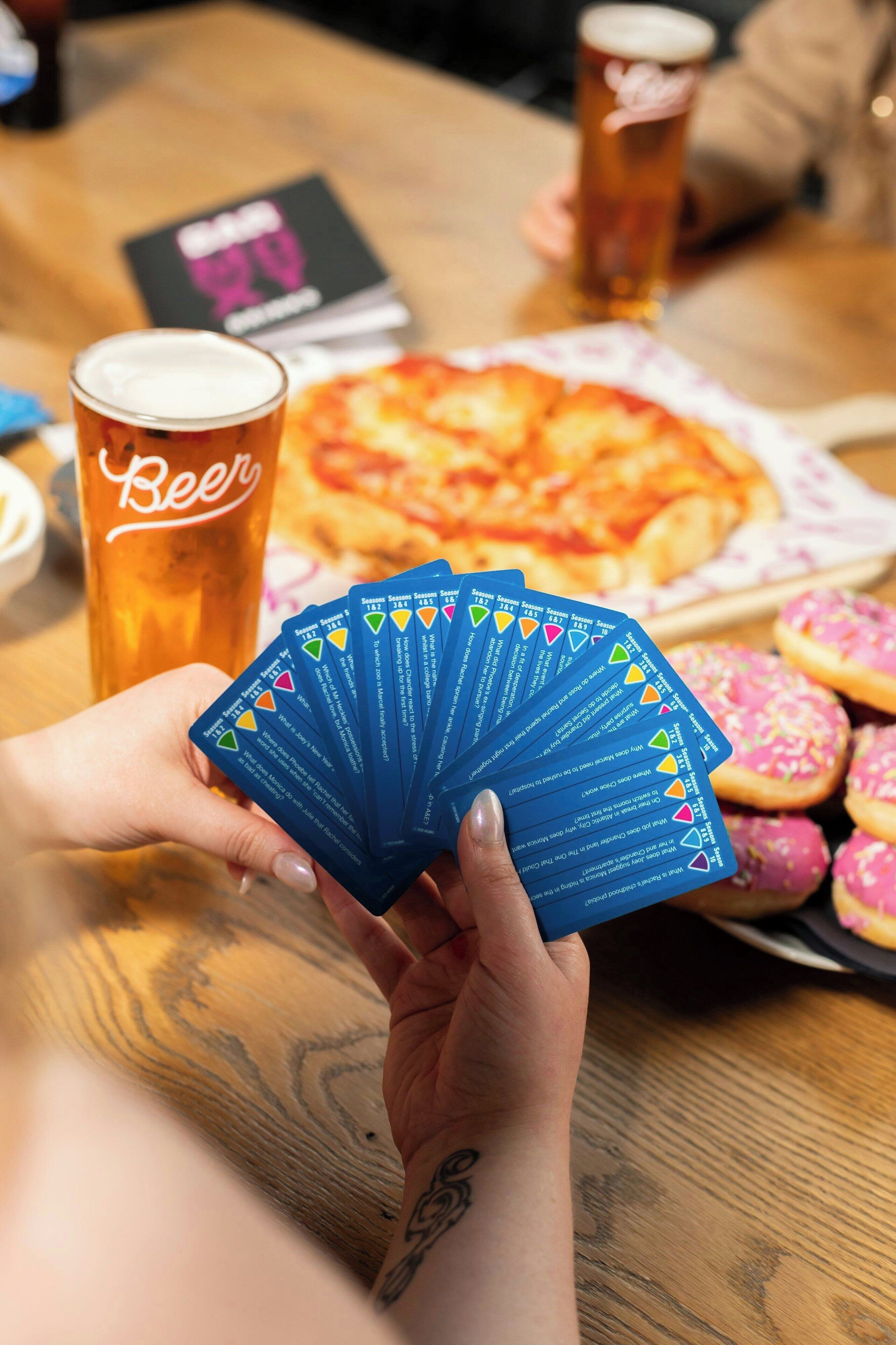 A person holding a deck of cards at a table with a pizza and a beer cup.