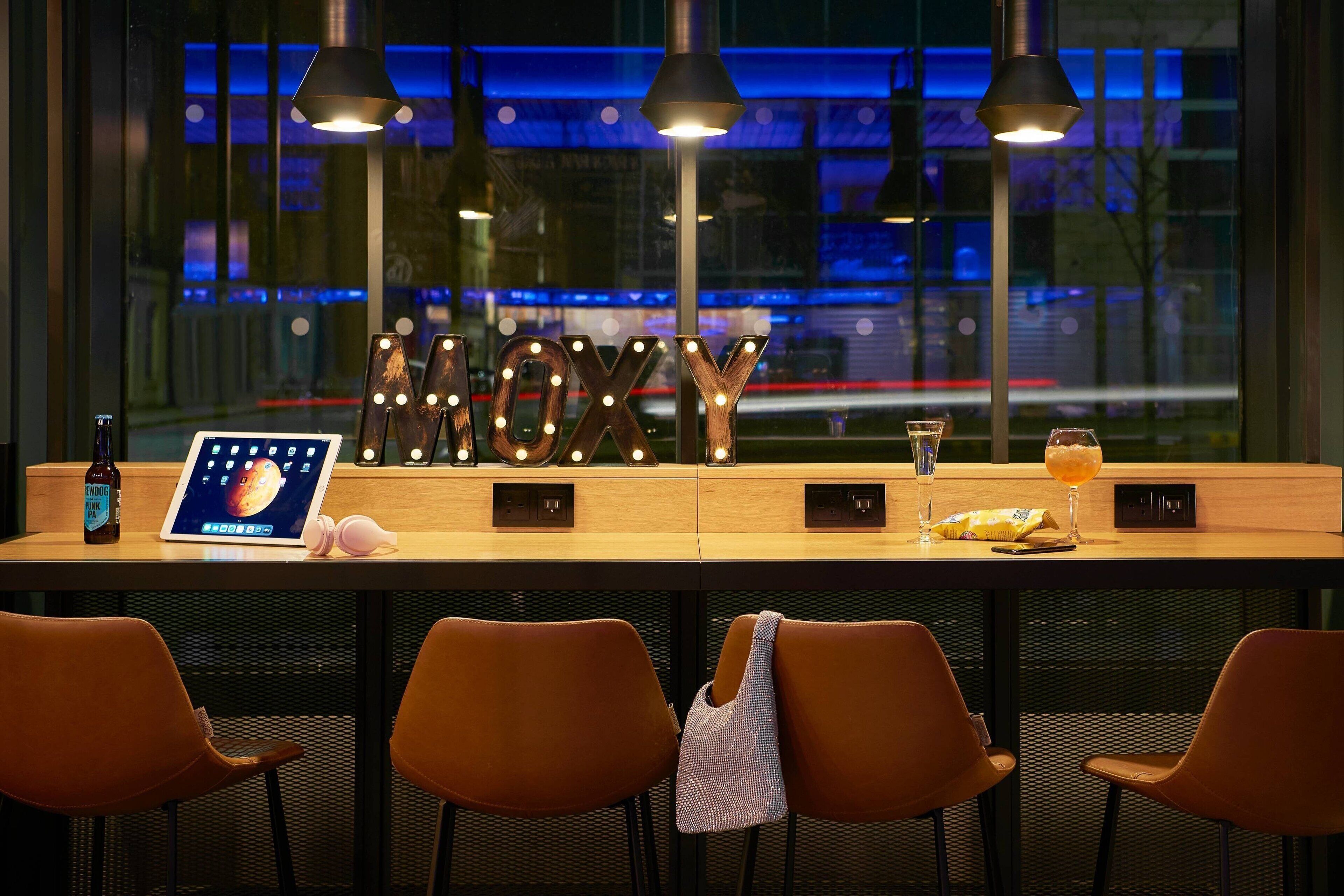 Een bar met een houten toonbank, verlichte letters die 'MOKY' spellen, een tablet, een biertje en glazen.