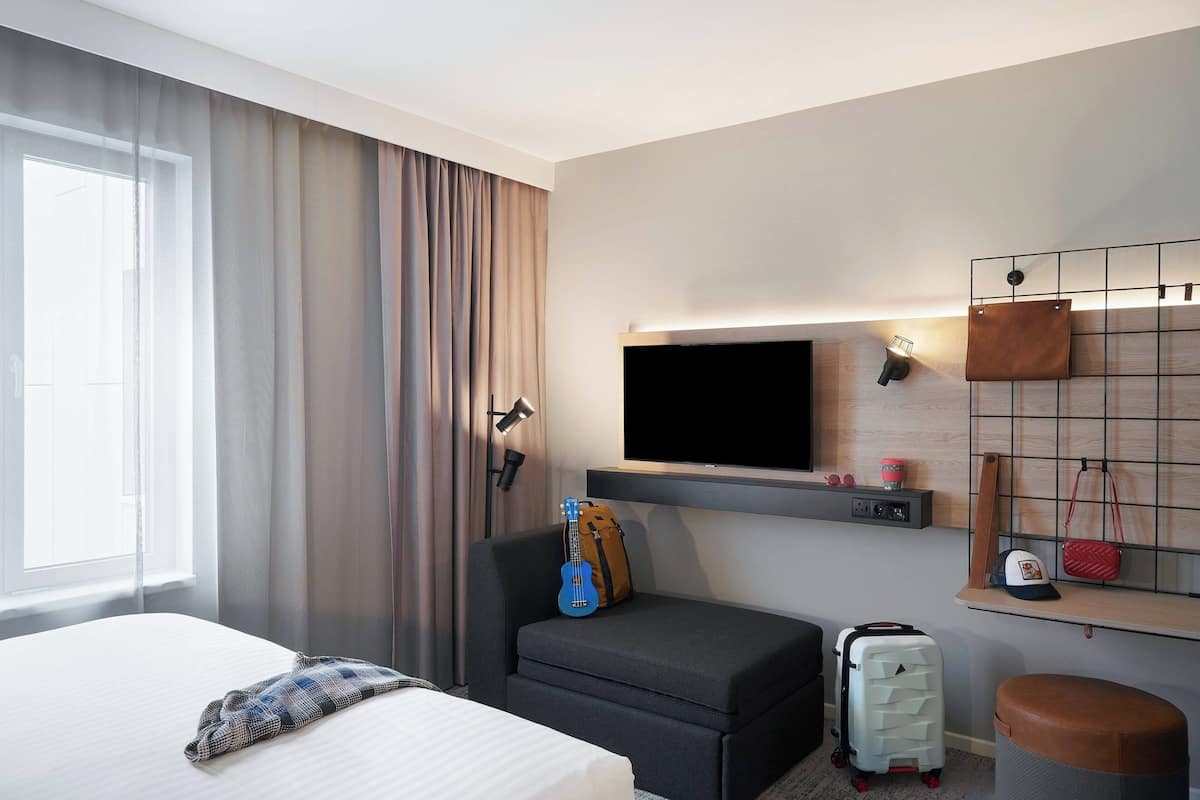 Une chambre d’hôtel avec un lit, un coin salon avec une guitare, un téléviseur fixé au mur, une valise et un petit tabouret.
