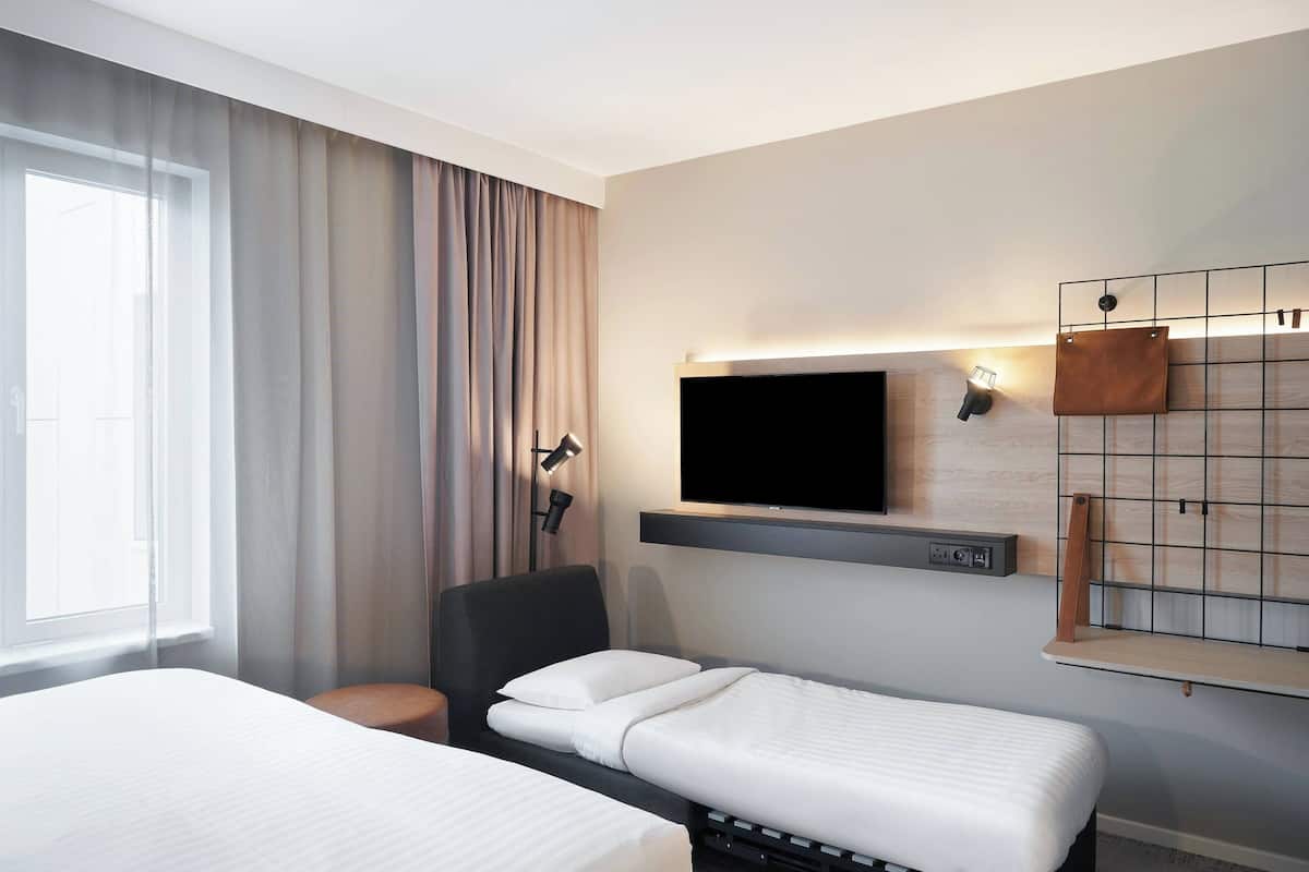 Une chambre d’hôtel avec deux lits, un téléviseur fixé au mur, un bureau avec une chaise et une fenêtre avec des rideaux.