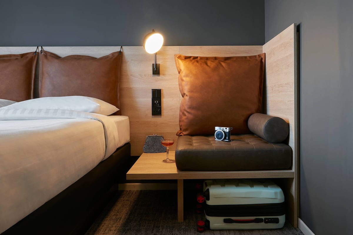 Une chambre d’hôtel avec un lit, une petite table sur laquelle se trouve un appareil photo, une lampe et une valise posée sous la table.
