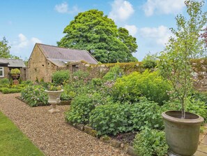 A stone building with a garden in front, a large tree, and a clear sky.