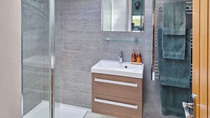 Une salle de bain moderne avec une douche à l’italienne en verre, un lavabo blanc avec deux flacons de savon et un meuble-lavabo en bois surmonté d’un miroir.