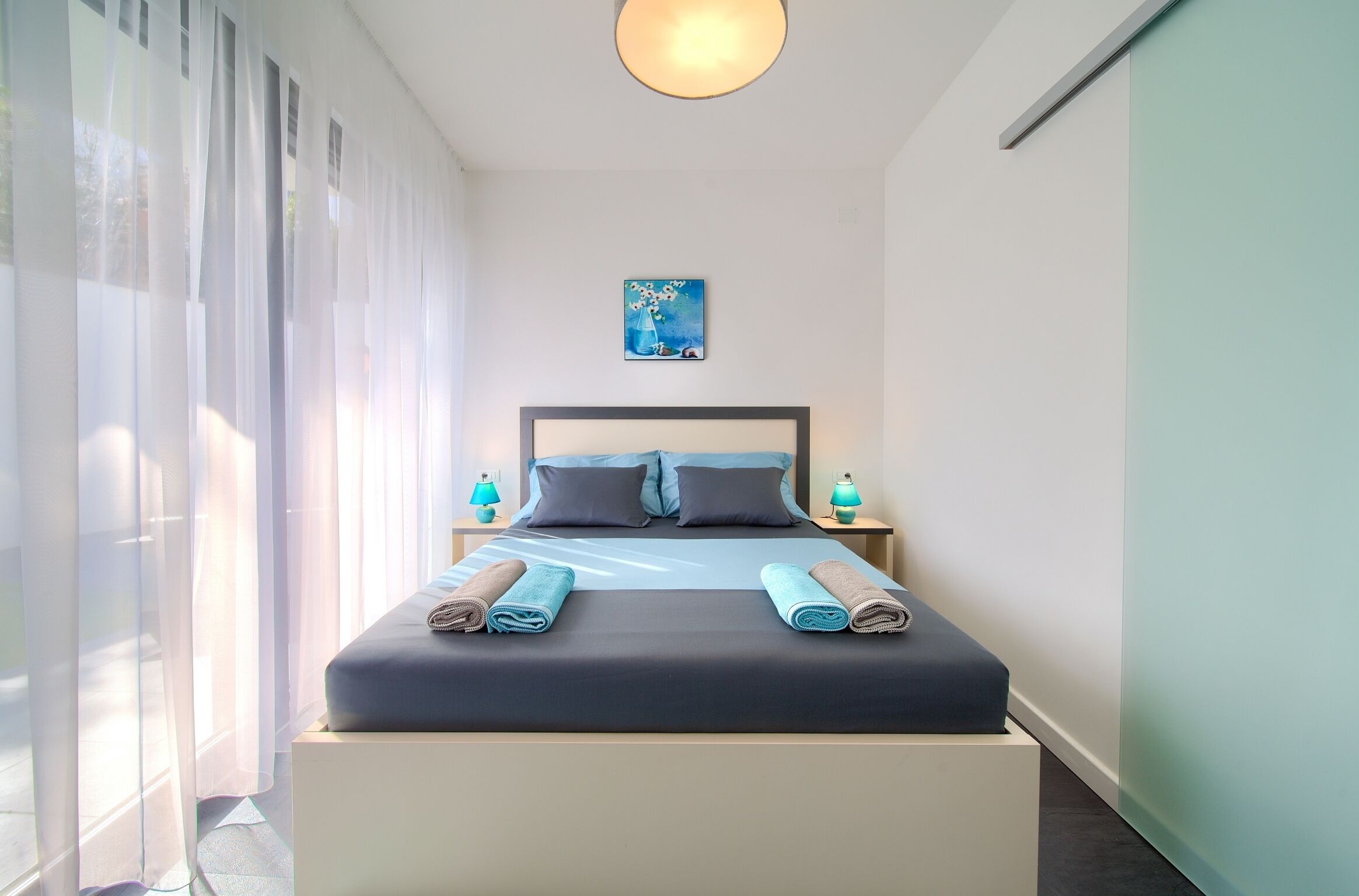 A neatly made bed with pillows and towels, a bedside table with a lamp, and a painting on the wall.