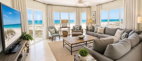 A spacious living room with a large flat-screen TV, comfortable seating, and a view of the beach.