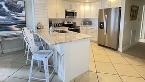 A modern kitchen with white cabinetry, a large island with bar stools, stainless steel appliances, and a view of the outdoors. - Emerald Isle 108 Beach Front First Floor End Unit (Pensacola Beach)