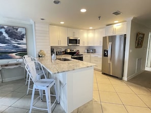 A modern kitchen with white cabinetry, a large island with bar stools, stainless steel appliances, and a view of the outdoors. - Emerald Isle 108, Beach Front, Sleeps 7- First Floor, End Unit w/ Balcony (Pensacola Beach)