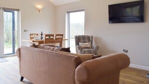 A living room with a sofa, a plaid armchair, a dining table with chairs, a TV mounted on the wall, and a large window. - 2 bedroom accommodation in Cwrt Newydd, near Llandysul (Cwrt Newydd, near Llandysul)