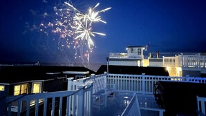 A rooftop with a white fence, a building with a flat roof, and fireworks in the background.
