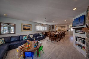 A spacious living area with a dark-colored sofa, a coffee table with children's toys, a dining area with multiple chairs, and a kitchen in the background.